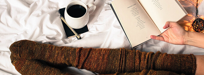 Frau in Strümpfen sitzt im Bett mit Buch, einem Notizbuch und Tee