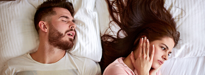 Frau hält sich die Ohren zu, während ihr Mann schnarcht.